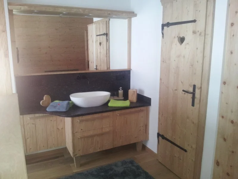 Badezimmer mit Holzregal, Waschtisch mit Waschbecken, Handtuch, Seifenspender, Holzdekoration und Holztür.