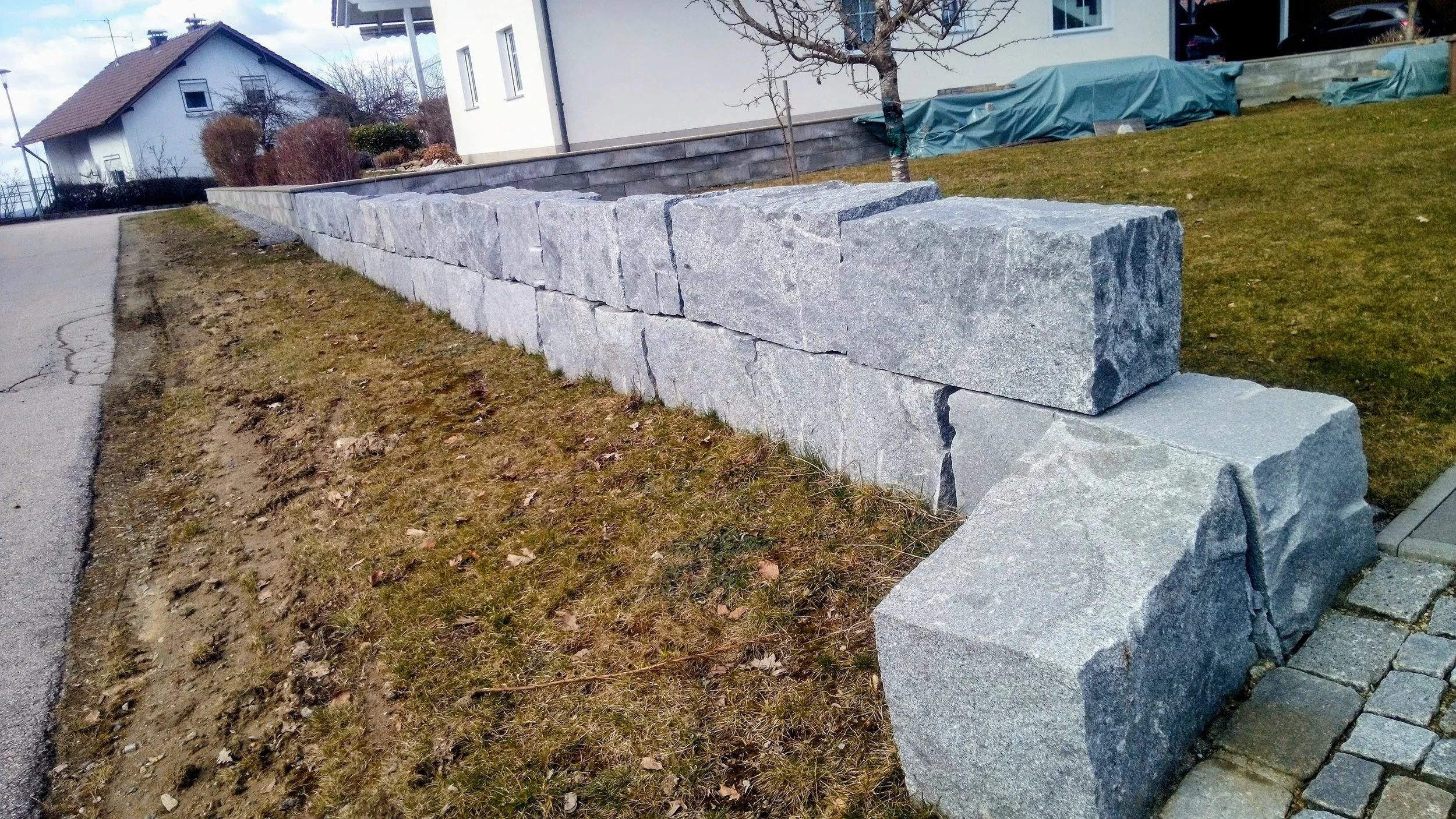 Reihe aus großen, unregelmäßig geformten grauen Steinen als Mauerspaltung entlang einer Gehwegseite. Im Hintergrund Häuser und Bäume.