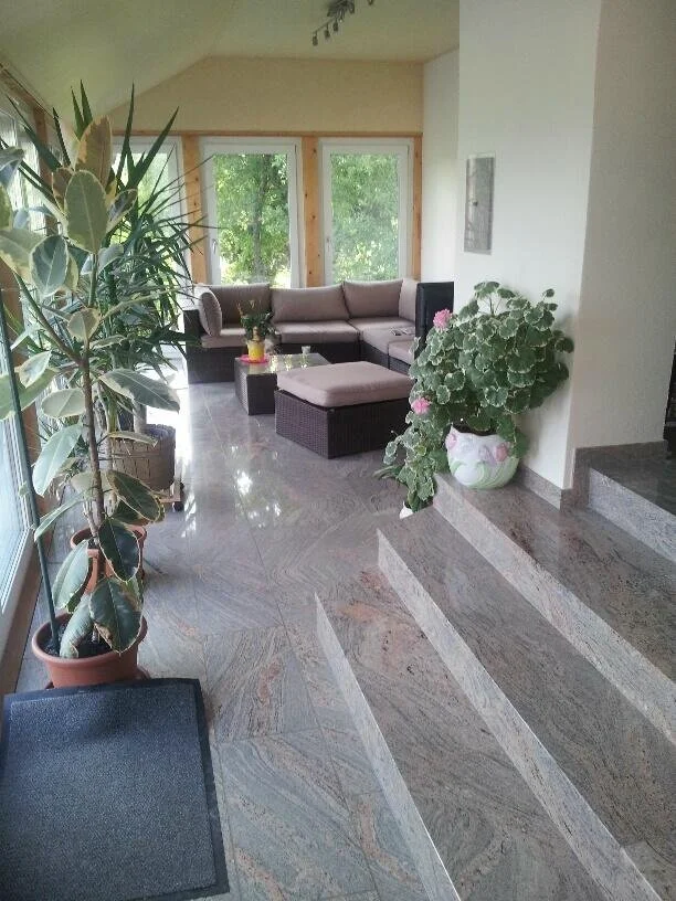 Gemütliches Wohnzimmer mit großen Fenstern, einer grauen Eckcouch, einem schwarzen Couchtisch, mehreren Pflanzen und einem hellen, einladenden Ambiente.