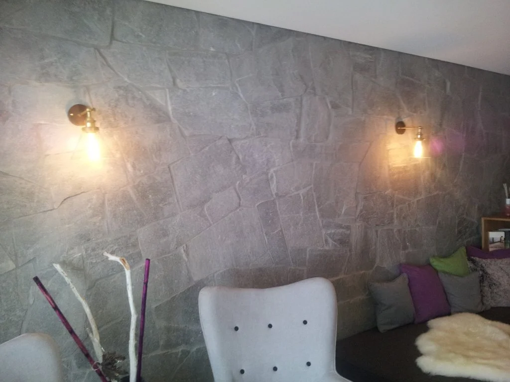 Wohnzimmer mit Steinwand, zwei Wandlampen, weißen Stühlen, Couch mit Kissen und Fell, kleiner Tisch mit Dekoration.