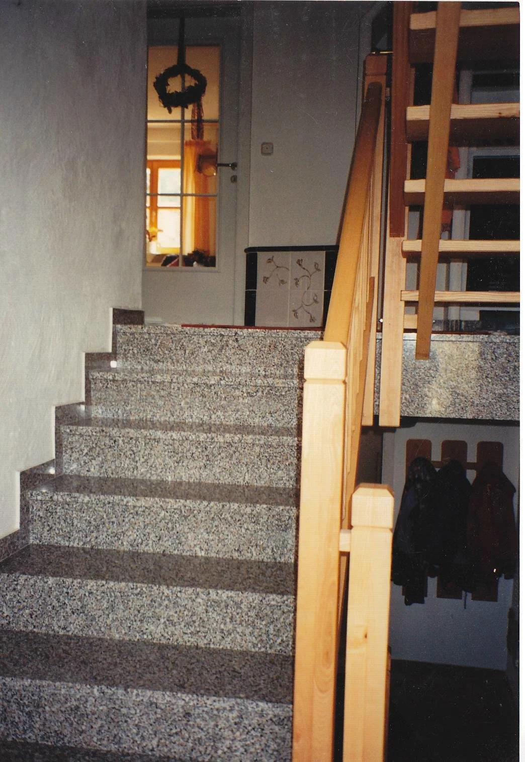 Innenaufnahme eines Hauses mit Treppe, Holzgeländer und Fliesenboden, Blick durch eine offene Tür mit Fenster auf einen sonnigen Raum mit bläulichem Himmel.
