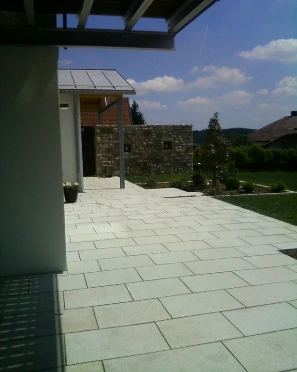 Ein moderner Terrassenbereich mit großen hellen Fliesen, ein Pflanzenkübel mit weißen Blumen, im Hintergrund eine Steinmauer und einige Bäume, unter einem Dachüberstand im Freien, bei sonnigem Himmel.