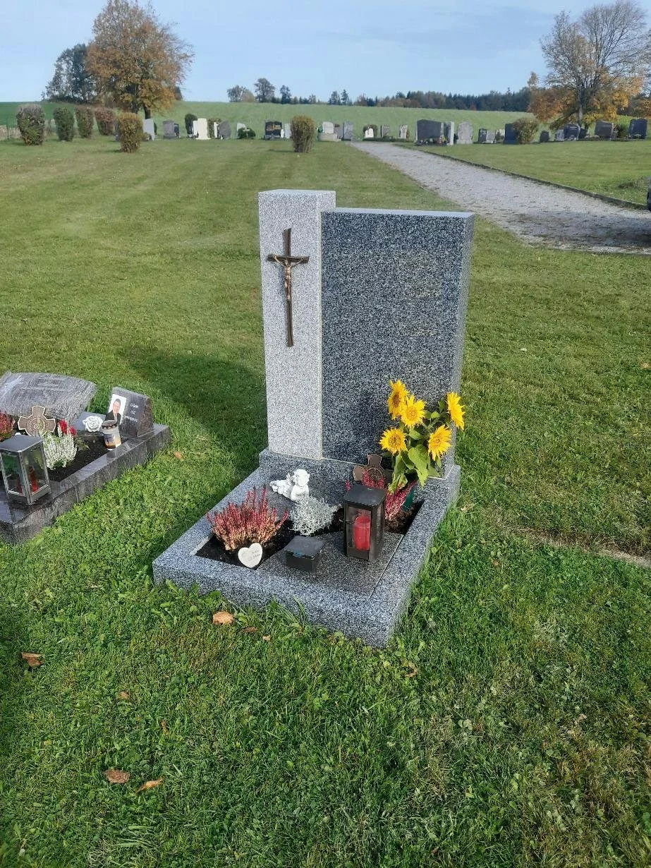 Ein Grab auf einem Friedhof mit einem Grabstein aus grauem Granit, einem kleinen Engel, Sonnenblumen, Kerzen und Dekorationen davor, umgeben von grüner Wiese und Bäumen im Hintergrund.