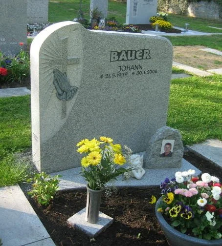 Granit-Gedenkstein für Johann Bauer mit einem Kreuz, einer Taube, einem Engel und einer Photografie, umgeben von Blumen und Kerzen im Friedhof.