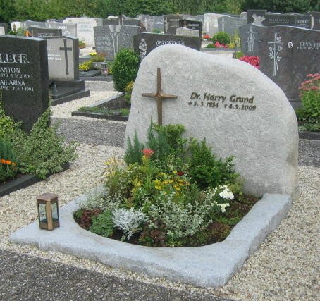 Gräber im Friedhof mit einem großen Stein, einem Kreuz und einem liebevoll gepflegten Grab mit Blumen und Kerze, Gedenktafel für Dr. Harry Grund.