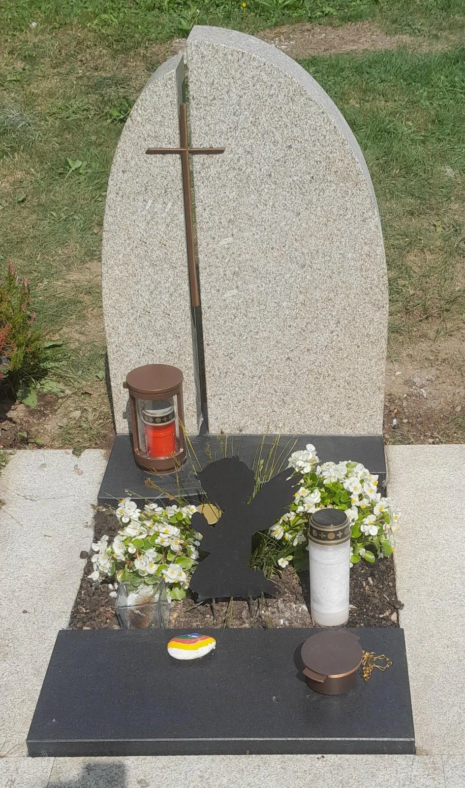 Granitgrabstein mit schwarzen Engelsfigur, Blumen, Laternen und einem Stein in Regenbogenfarben, auf einem Friedhof