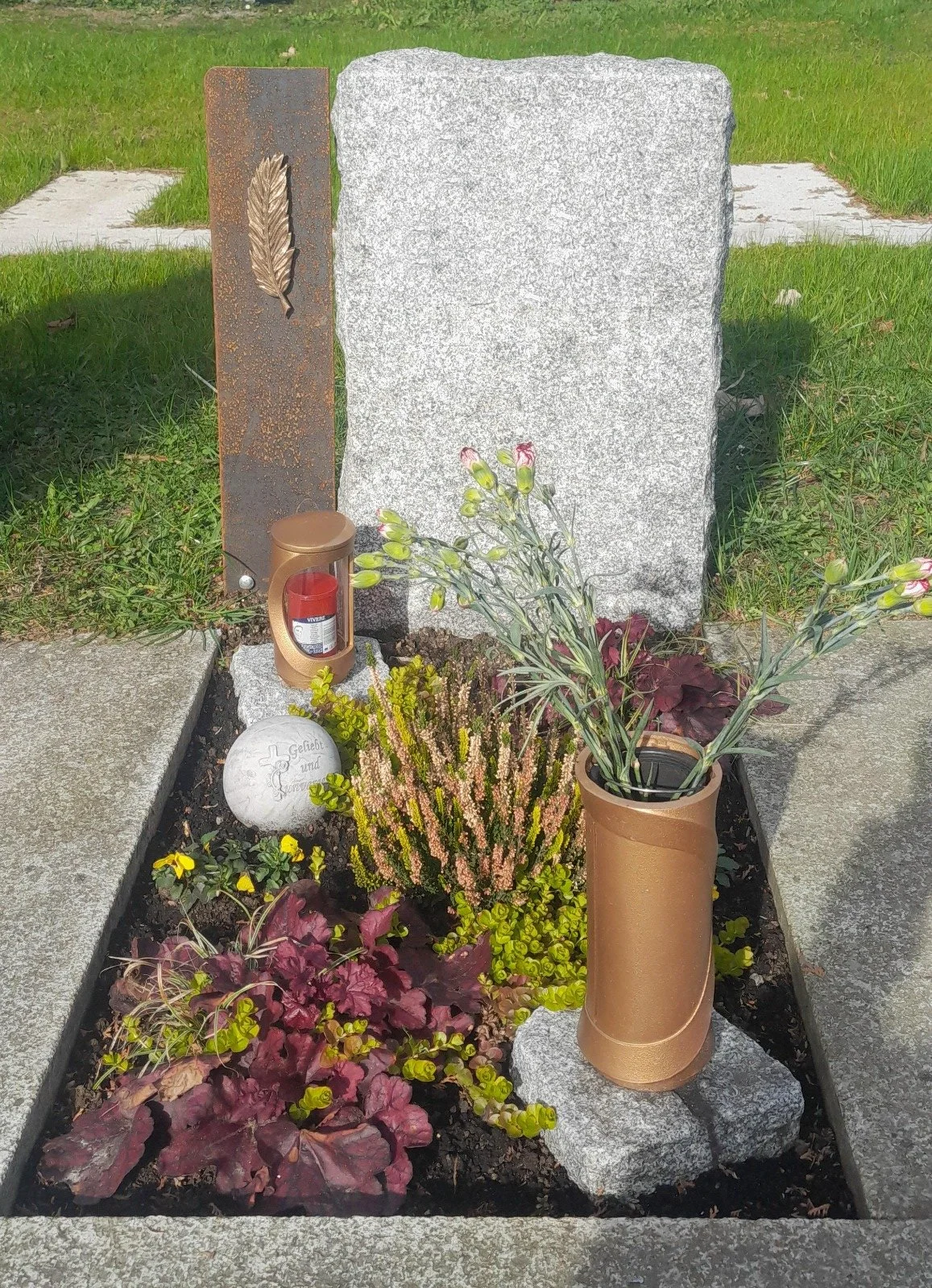 Ein Grabschmuck mit Blumen, Gedenkenkerze, Stein mit belegter Gedenkplatte, auf Friedhof.
