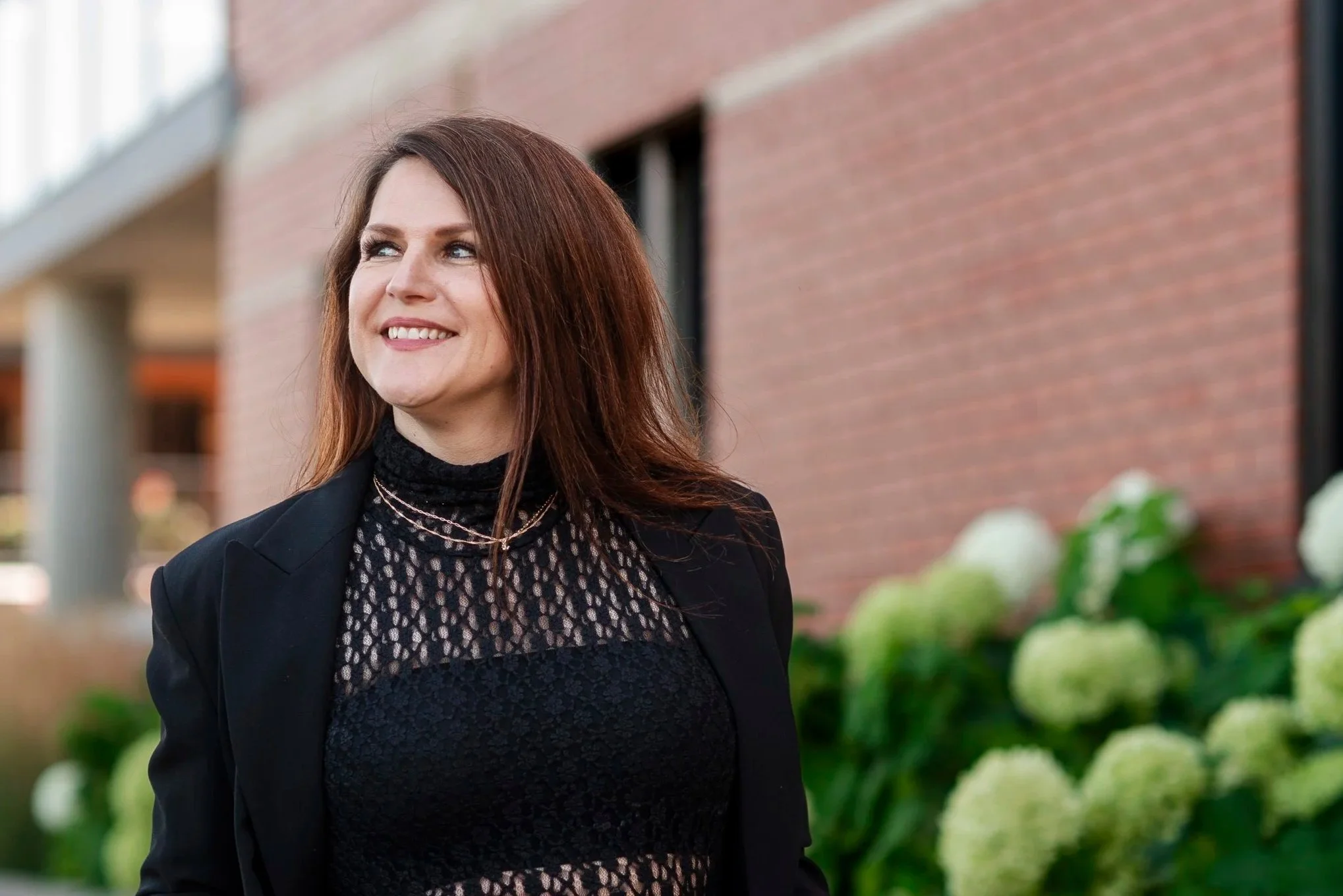 A woman with long brown hair wearing a black blazer and black lace top standing outdoors near a brick building with bushes in the foreground, smiling and looking slightly to her right.