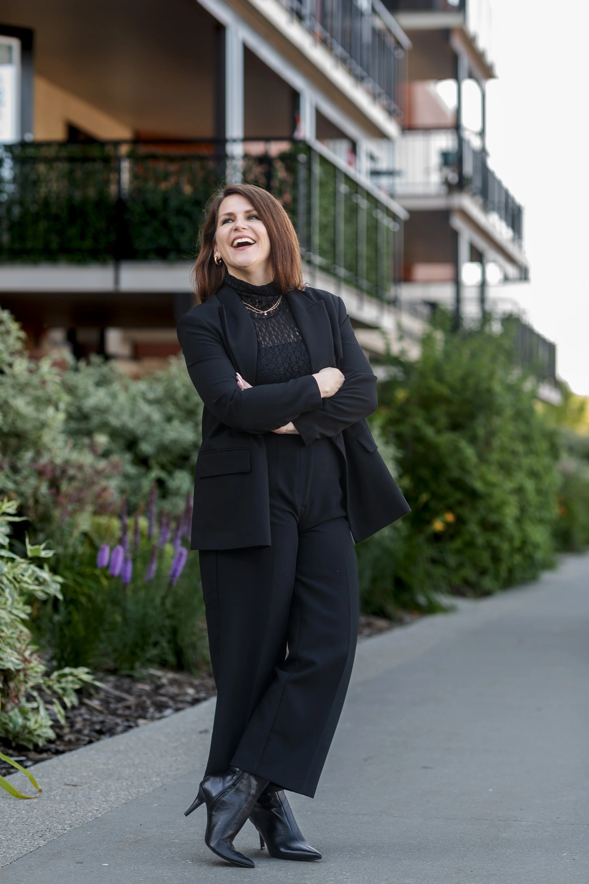 A woman standing outdoors on a sidewalk, dressed in a black suit with black high-heeled boots, smiling and crossing her arms, with a modern apartment building and greenery in the background.