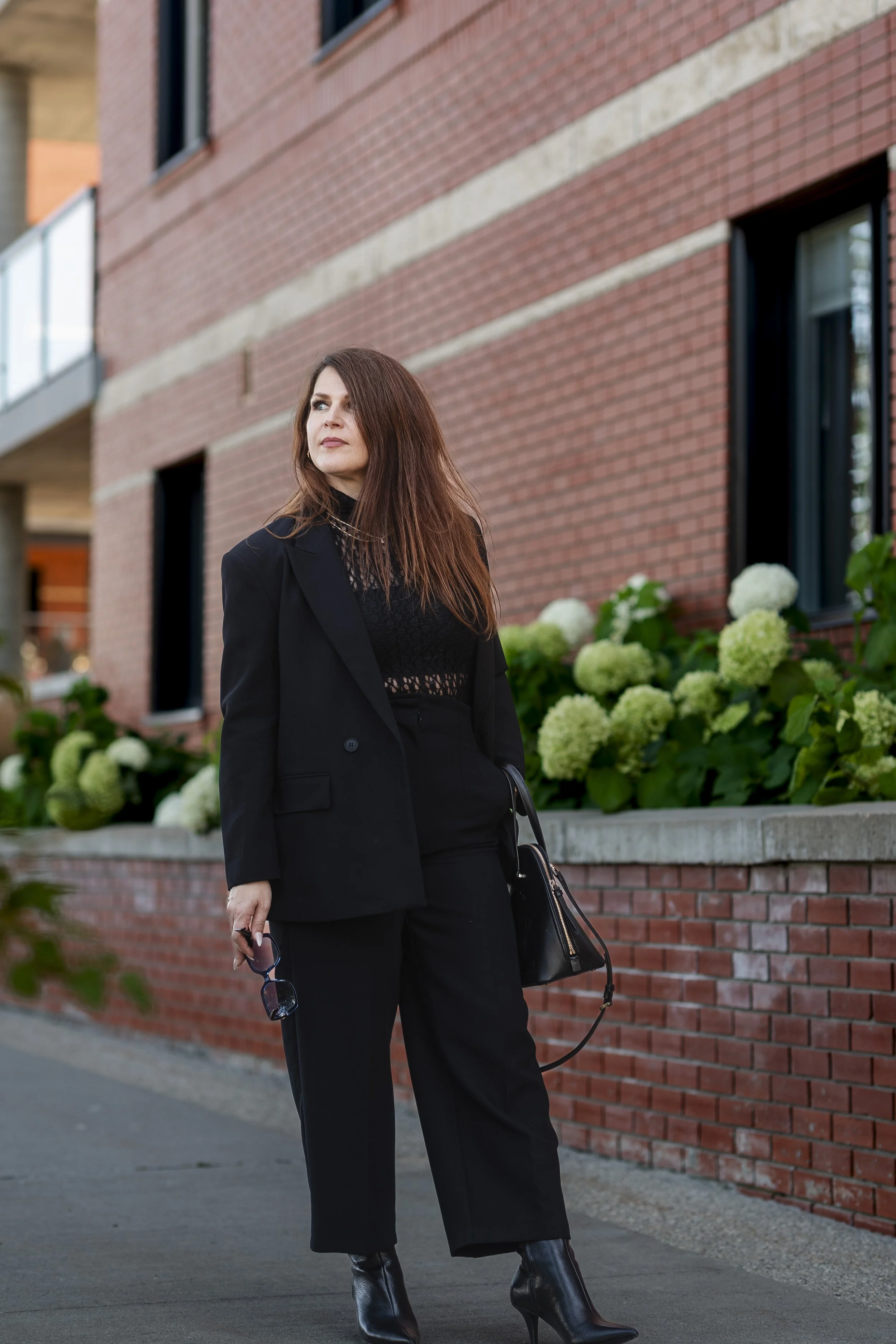 A woman standing outdoors in front of a brick building with large windows and white hydrangea flowers. She has long, reddish-brown hair and is wearing a black blazer, black pants, and black heeled boots. She is holding sunglasses in her right hand and a black handbag is hanging from her left shoulder.