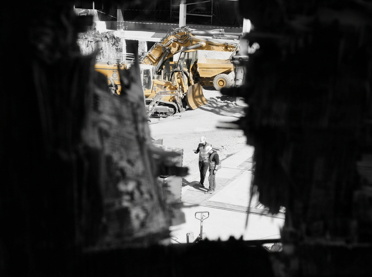Photo taken through a dark, torn opening showing a construction site with two workers, a yellow excavator, and building structures in the background.