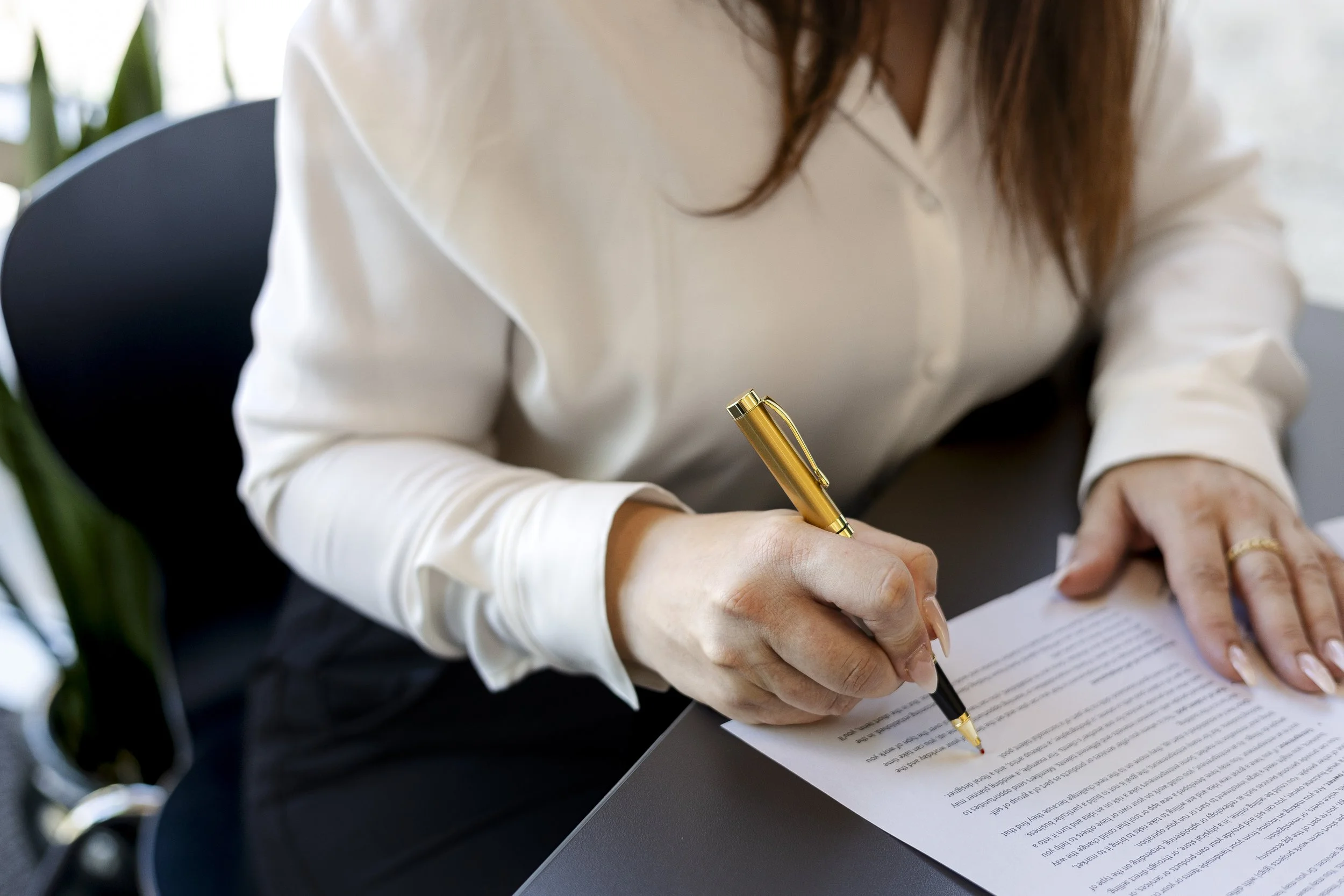 A woman with brown hair wearing a white shirt is sitting at a desk, signing a document with a gold and black pen.