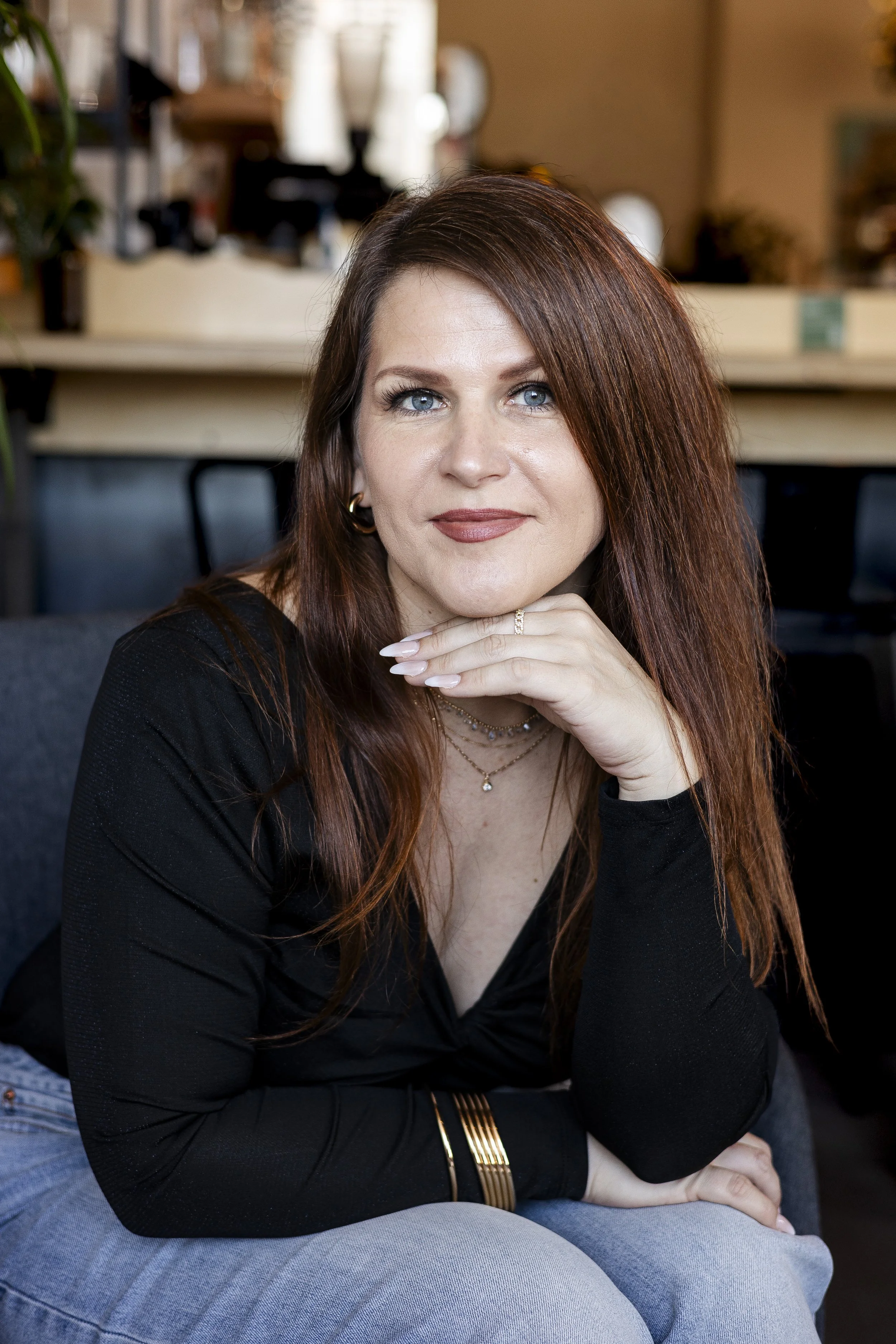 A woman with long, wavy red hair and blue eyes wearing a black top and layered jewelry, sitting on a gray couch in a cozy indoor space.