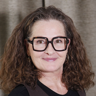 Kvinnor med brunt, flätat hår och stora, svarta glasögon som ler mot kameran, mot en beige gardin.