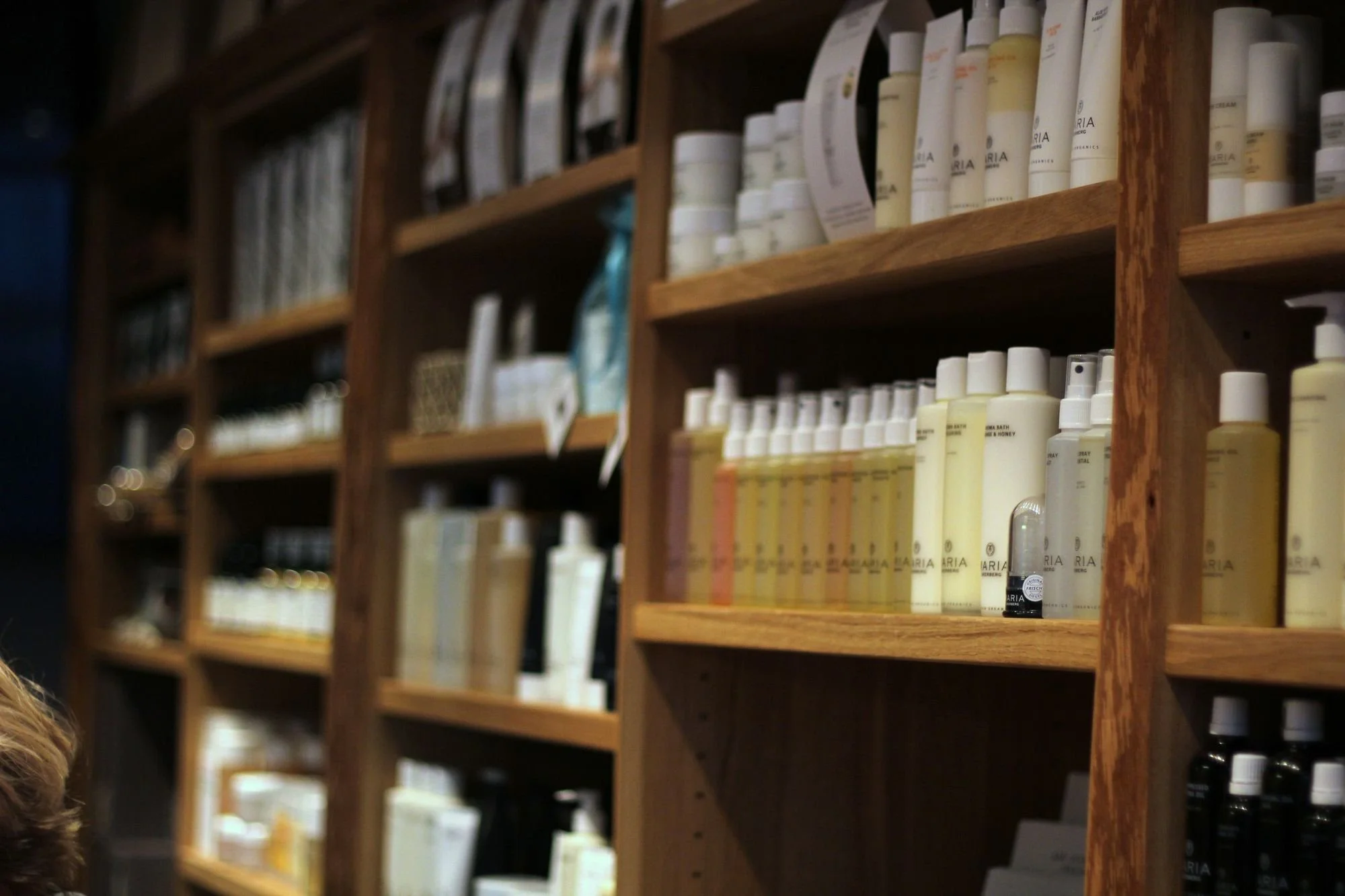 Bild av en trähylla fylld med vita och mörka flaskor och tuber, troligtvis skönhets- eller hudvårdsprodukter, i en butik.
