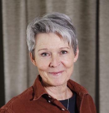 Porträtt av en äldre kvinna med kort, grått hår, klädd i en brun skjorta, mot en bakgrund av gardiner.