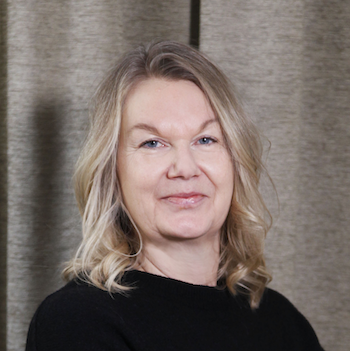 Porträtt av en lätt skägglös kvinna med blont, vackert lockigt hår, som ler svagt, iförd svart tröja och står mot en grå bakgrund.