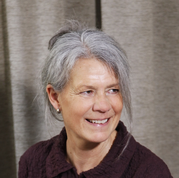 En äldre kvinna med grått hår, ler och tittar åt sidan, klädd i en mörk lila kofta.