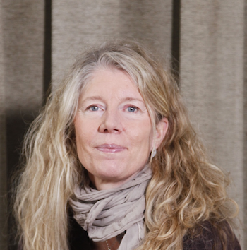 En kvinna med långt, lockigt blont hår, blå ögon och en ljus scarf runt halsen står framför en beige gardin.