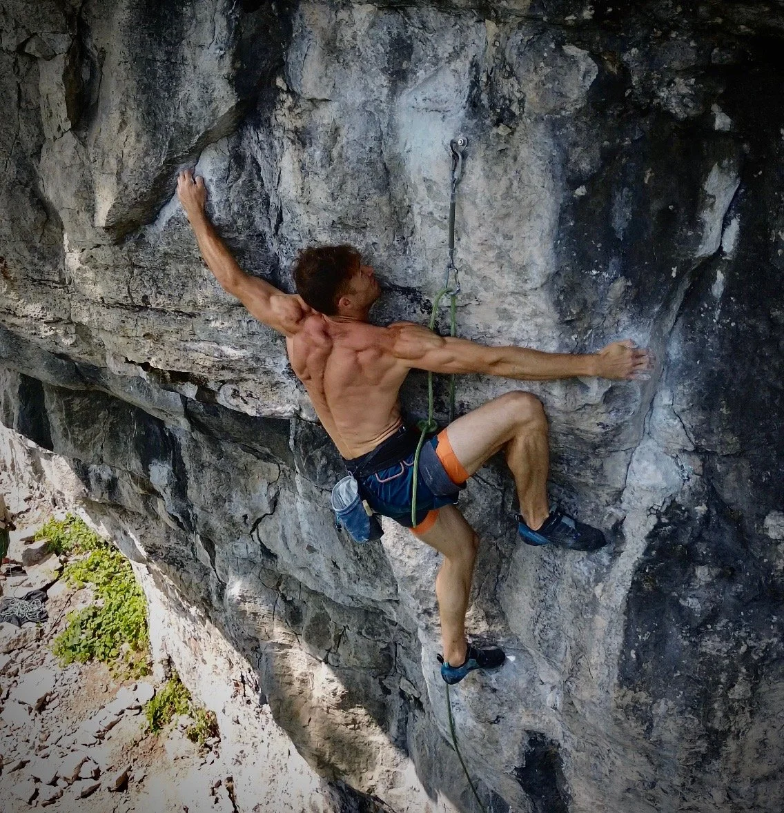 A male rock climber scaling a steep outdoor rock face, wearing climbing shoes and shorts, attached to a climbing rope for safety.