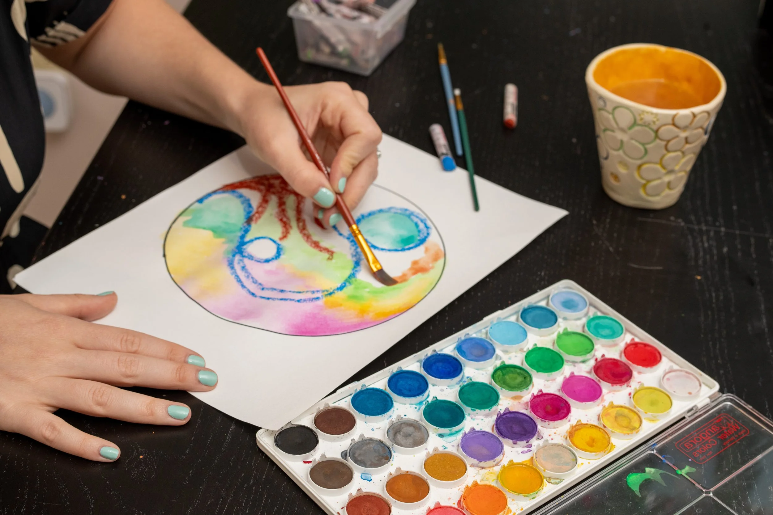 Person painting a colorful abstract design on a paper with watercolors, watercolor palette, and paintbrushes on a black table.