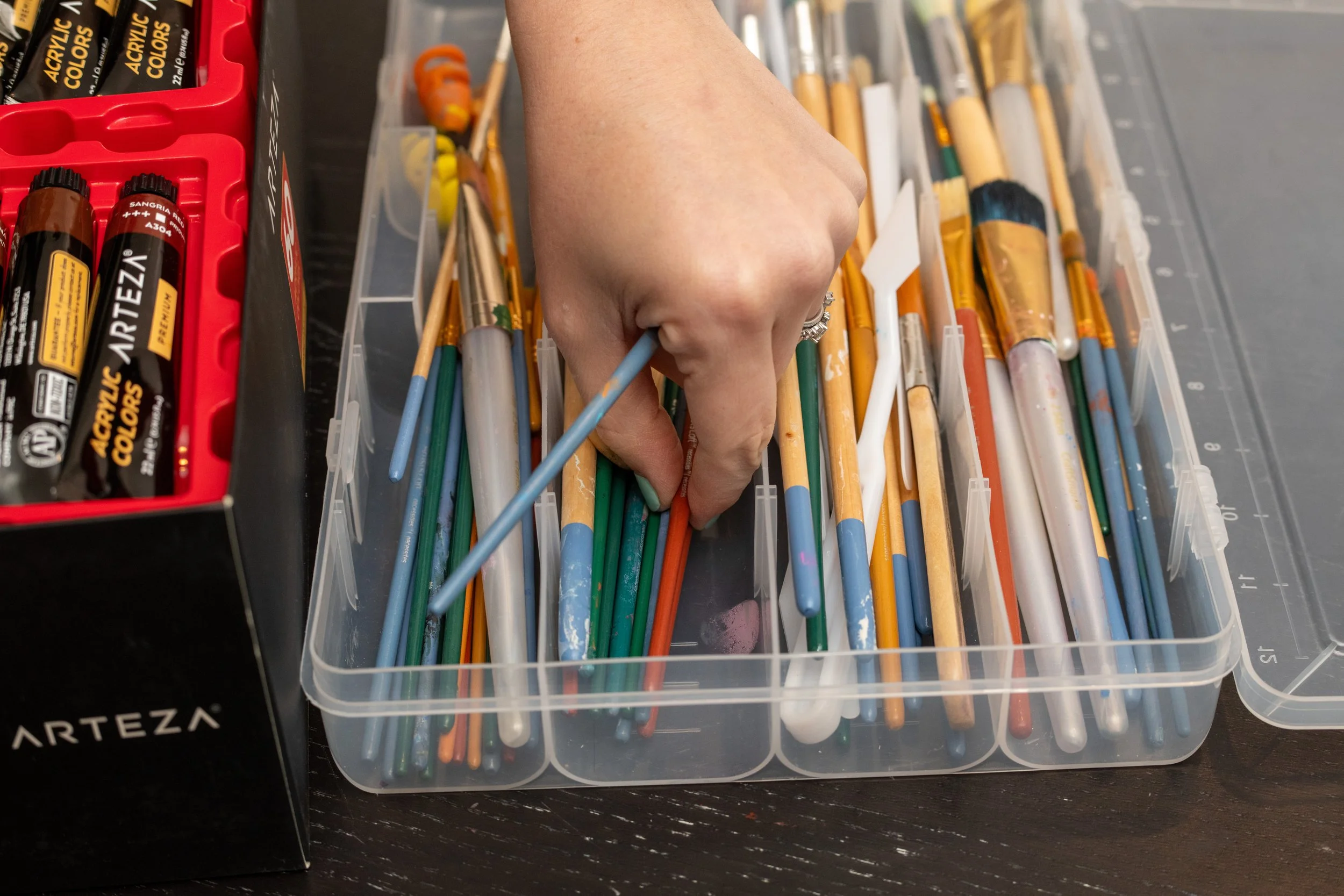 A hand sorting through a plastic container filled with colorful paintbrushes on a dark wooden table, with a box of acrylic paint sticks nearby.