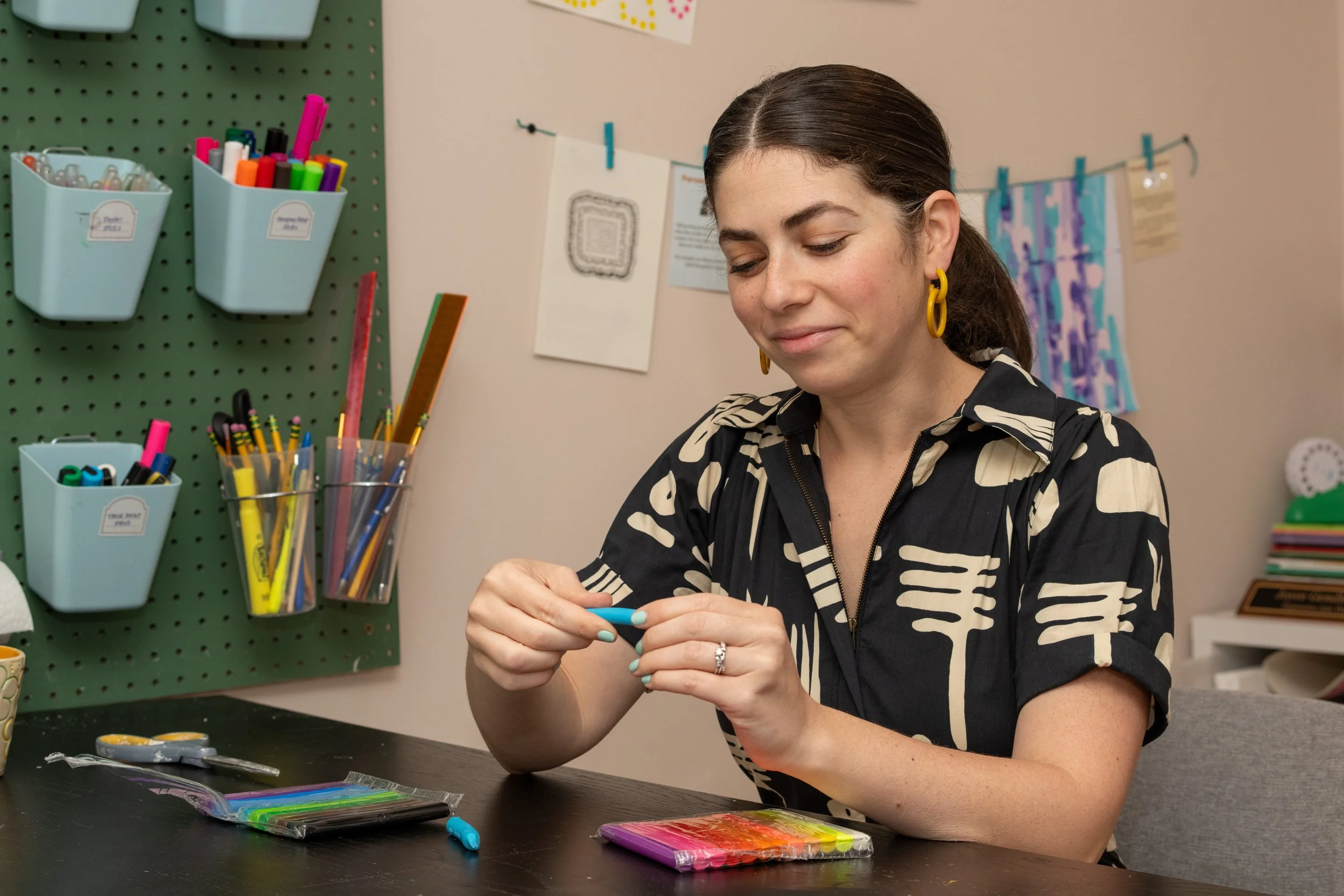 A woman with dark hair in a ponytail, wearing a black and beige patterned shirt and yellow hoop earrings, sitting at a desk and holding a small colorful object. The desk has various craft supplies, including packs of modeling clay, on it. The background features a pegboard with storage bins filled with supplies and a wall decorated with artwork and photos.