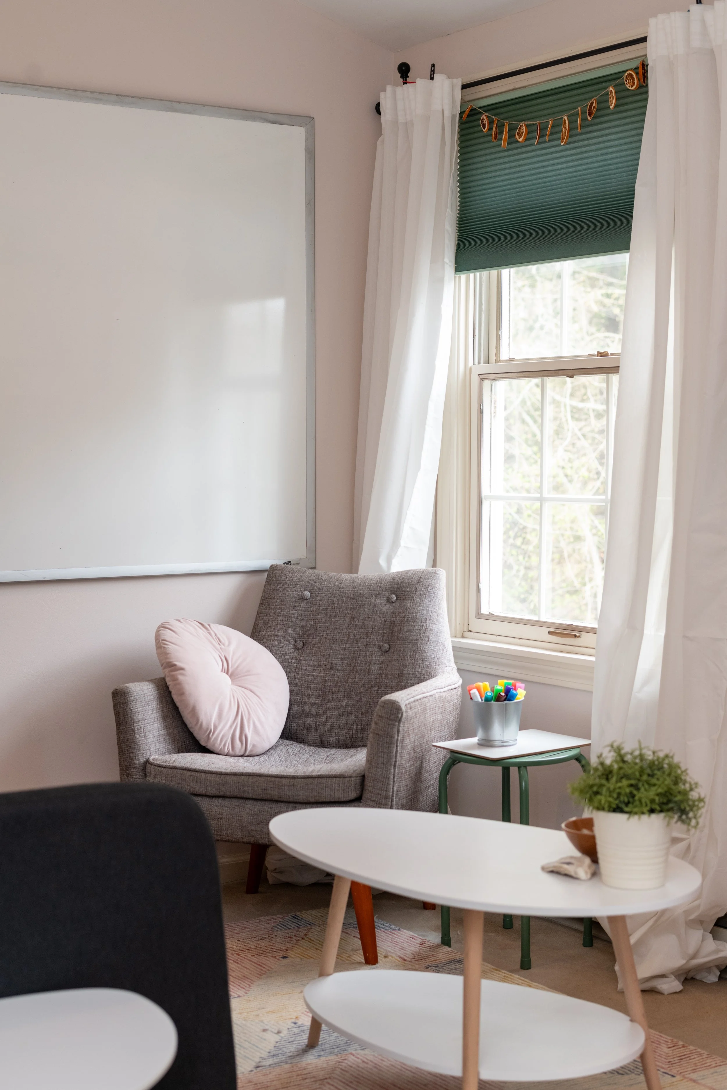 A cozy corner of a room with a gray armchair, pink cushion, a small side table with colorful markers, a white oval table with a potted plant and ceramic bowl, a window with white curtains, and a large whiteboard on the wall.