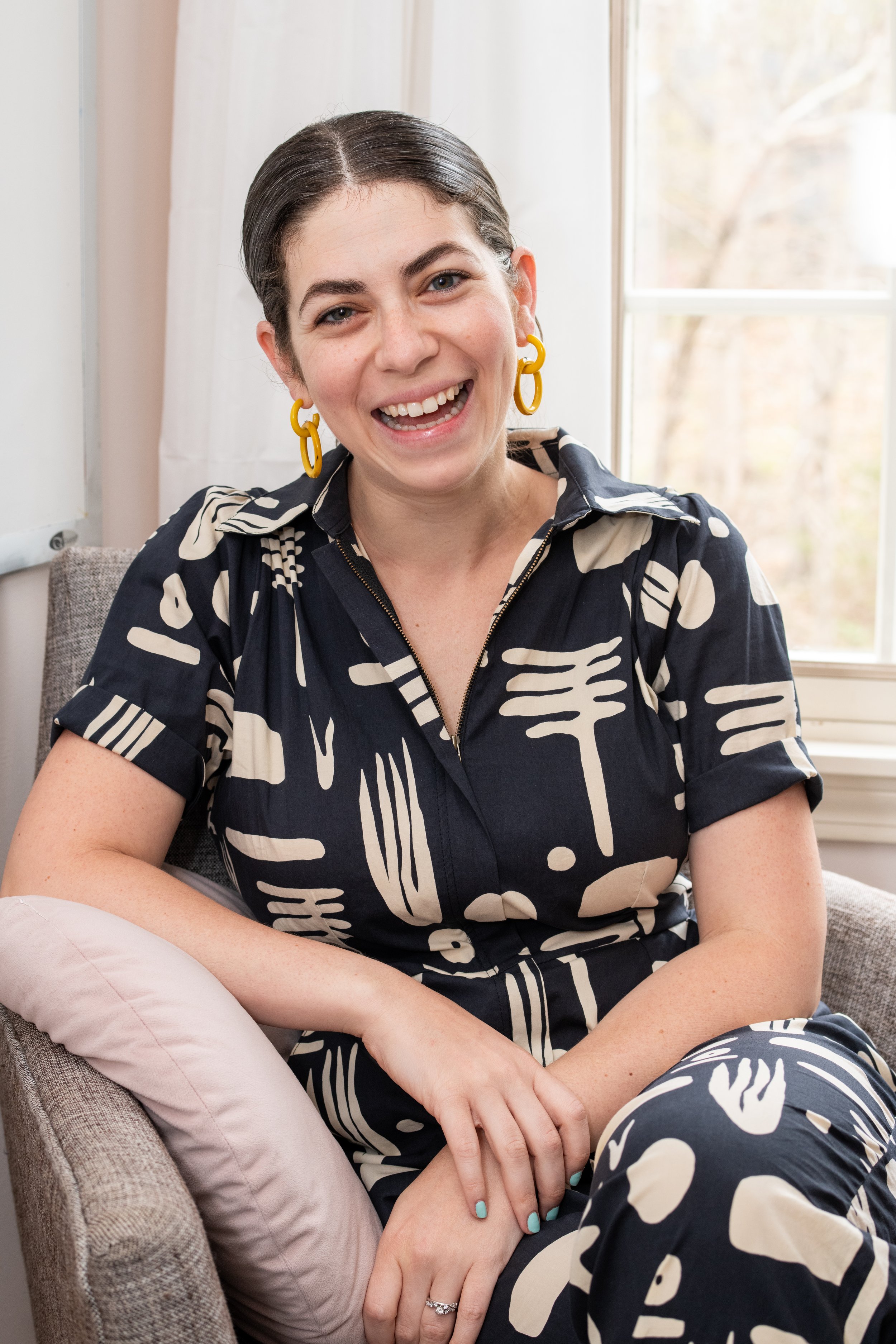 A woman with dark hair and yellow earrings sitting on a beige chair, smiling, in front of a window with a view of trees.