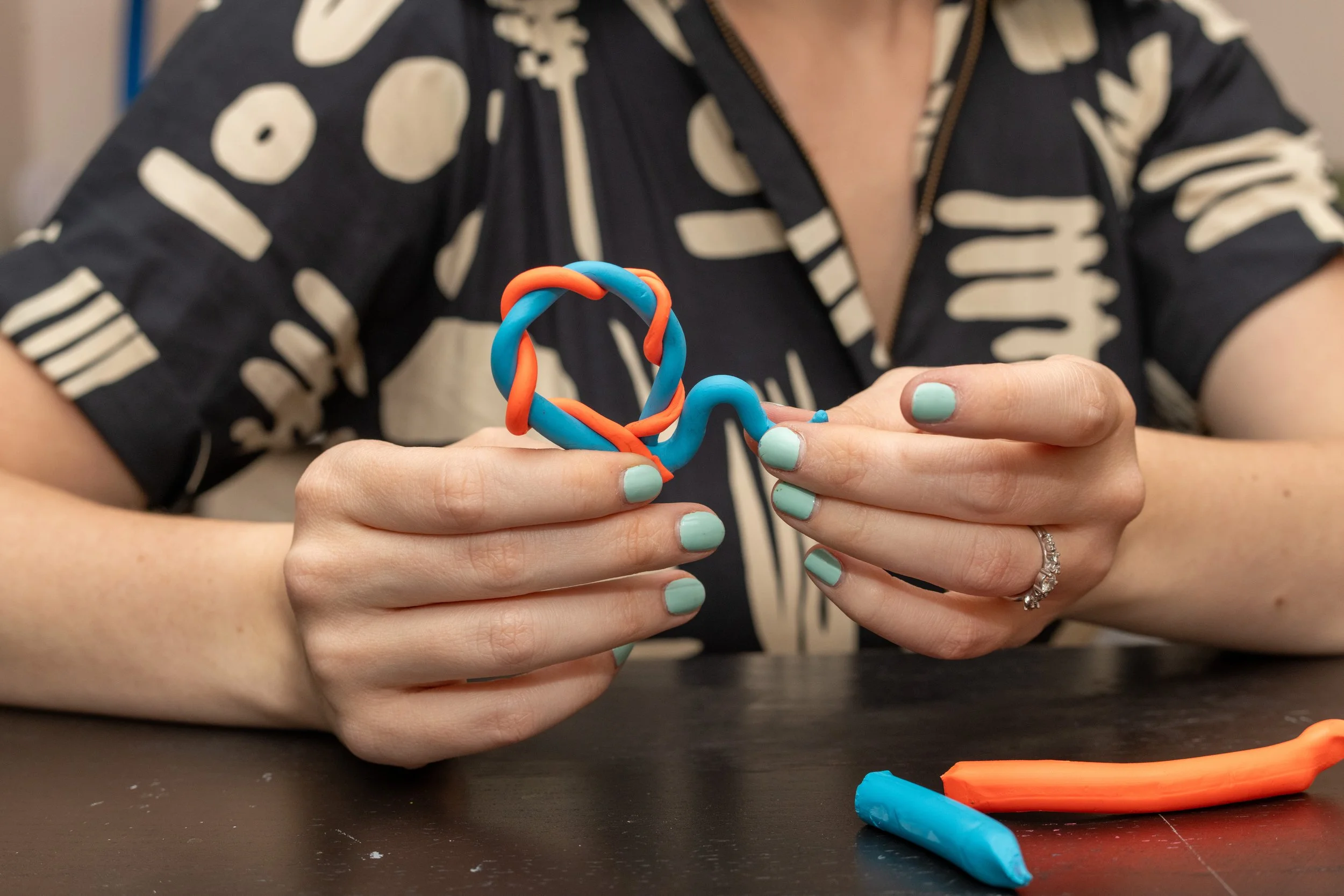 A woman with light green painted nails, wearing a ring on her left ring finger, is holding a colorful, twisted clay sculpture made of blue and orange clay. There are additional pieces of blue and orange clay on the table in front of her. She is wearing a short-sleeved black shirt with a white abstract pattern.