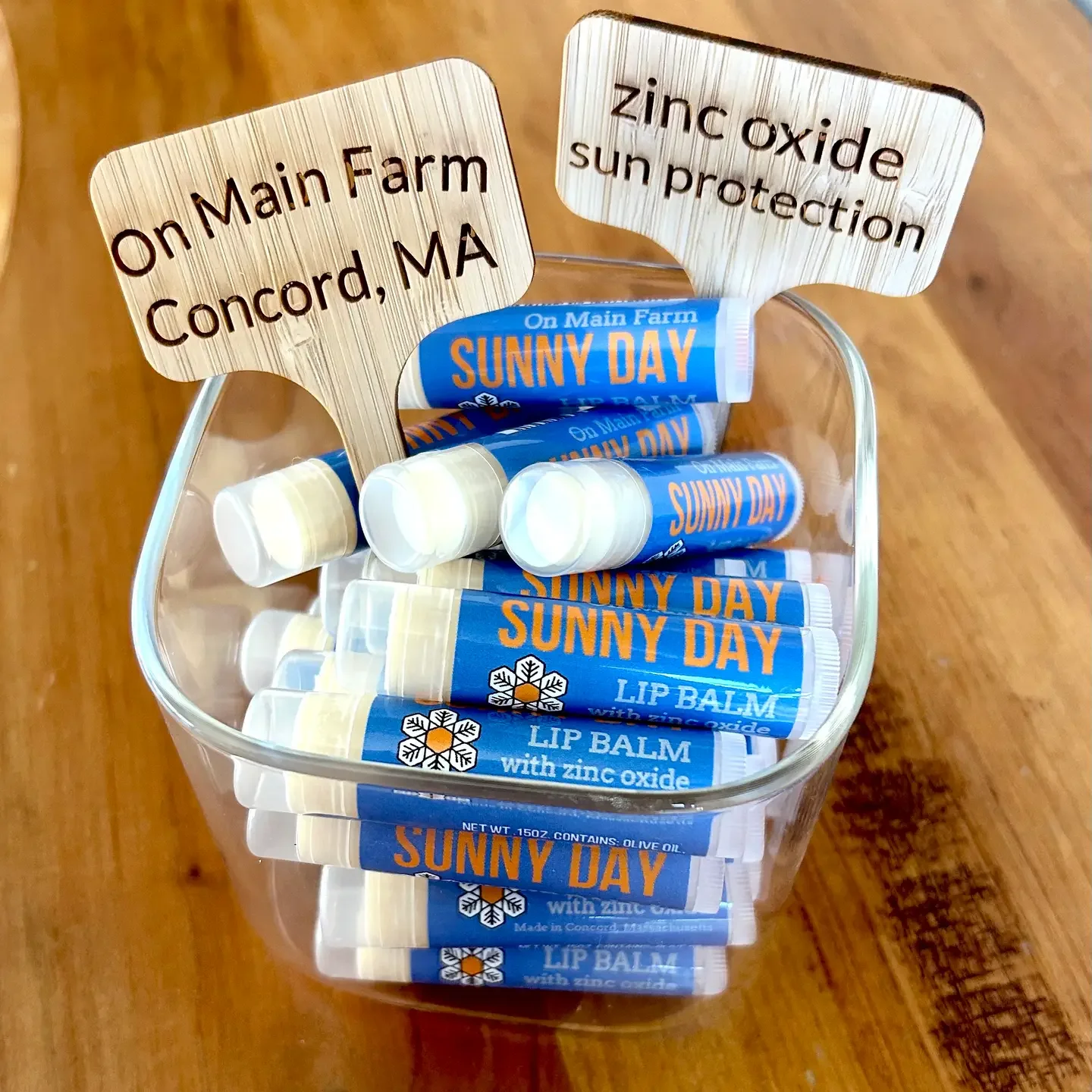 A clear plastic container filled with multiple tubes of Sunny Day lip balm with zinc oxide, placed on a wooden surface. Two wooden signs in the container contain the phrases "On Main Farm Concord, MA" and "zinc oxide sun protection."