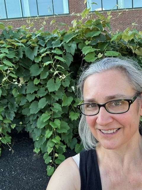 Bonnie Albright with gray hair and glasses smiling outdoors in front of green leafy plants.