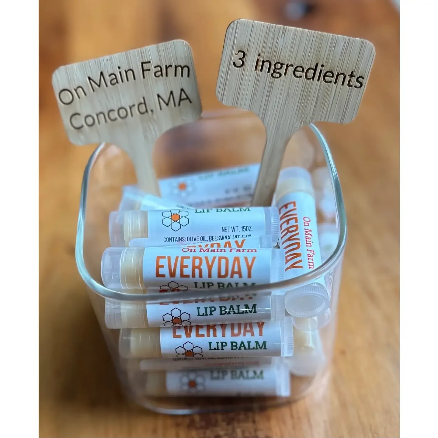 Small transparent container filled with several tubes of On Main Farm Everyday Lip Balm, with wooden signs stating "On Main Farm Concord, MA" and "3 ingredients" inserted into the container.