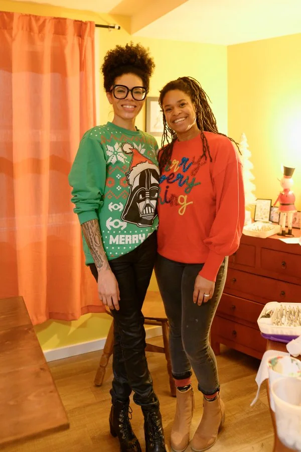 Two women smiling and standing inside a warmly lit room with yellow walls, pink curtains, and a wooden dresser with holiday decorations.