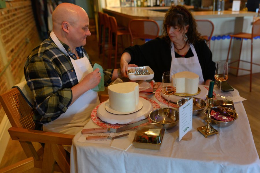 Two people sit at a table in a restaurant, decorating two white frosted cakes. The man wears a plaid shirt and the woman has dark curly hair and is wearing a black top. There are glasses of wine, decorating supplies, and a menu on the table.