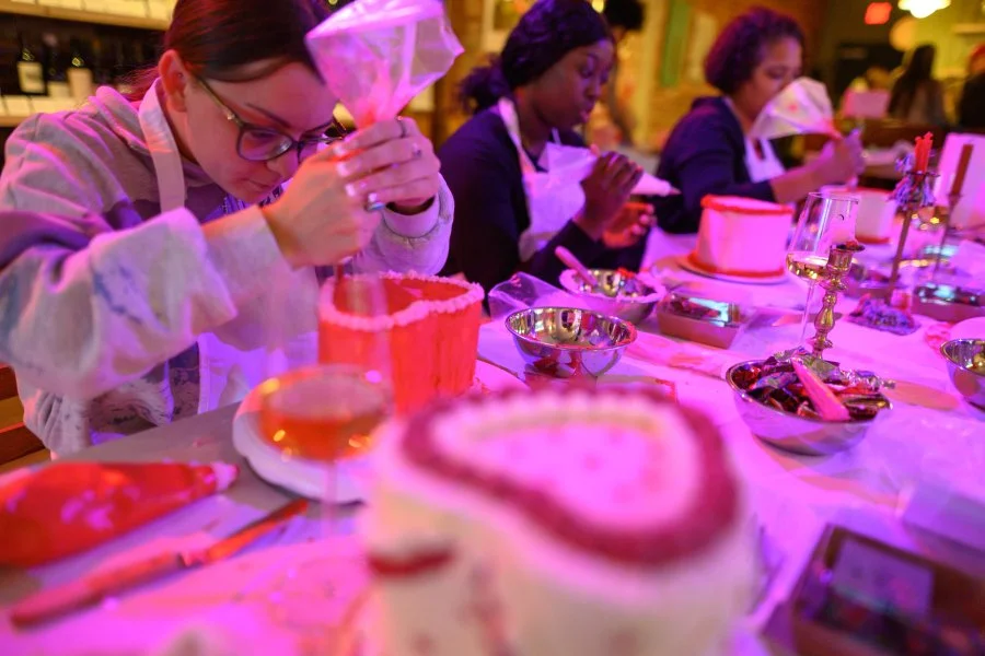 People gathered at a table decorating and preparing cakes and desserts for a celebration in a warmly lit indoor setting.