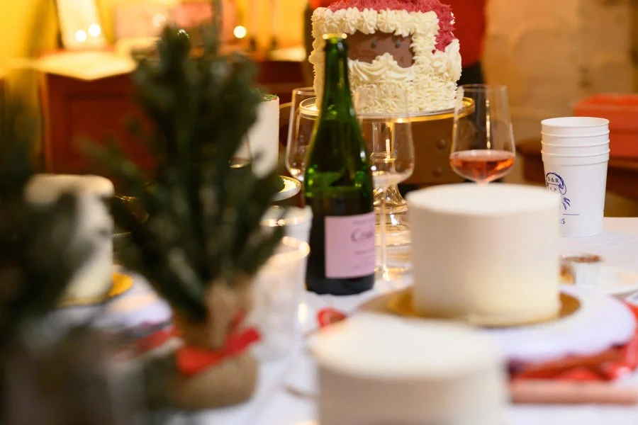 A table set for a celebration with cakes, drinks, and a small Christmas tree. In the background, a festive cake with white and red frosting and a photo insert is visible.