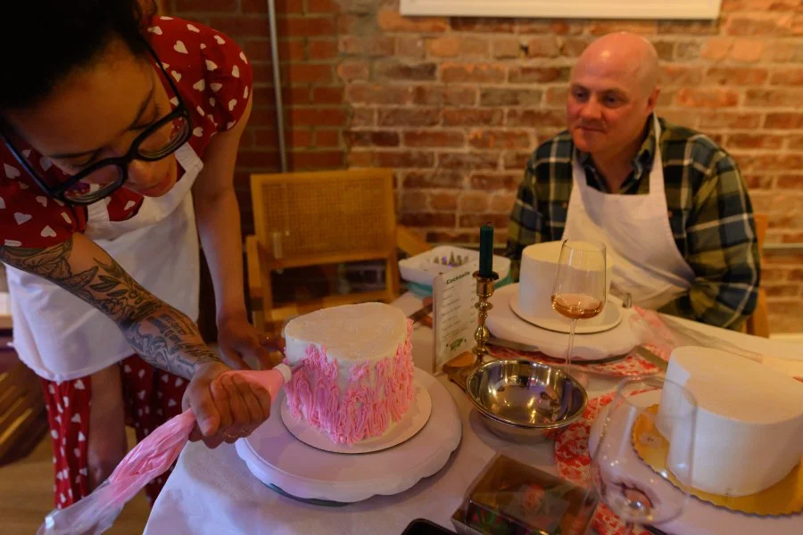 A woman in a red dress with white hearts cuts a pink and white cake, while a man in a plaid shirt with an apron observes at a table with a wine glass, a candle, and other dining items in a brick-walled room.