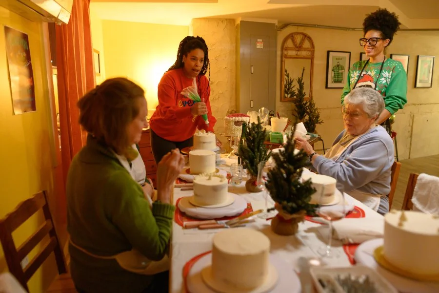 Family celebrating Christmas with cakes at a decorated dinner table, including young women and children, in a warmly lit room with holiday decorations.