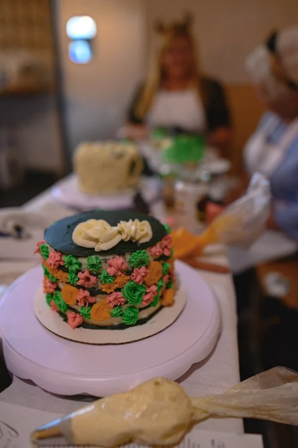 Colorful decorated cake with pink, green, and orange piping, topped with white frosting, on a pink cake stand in a room with people and another cake in the background.