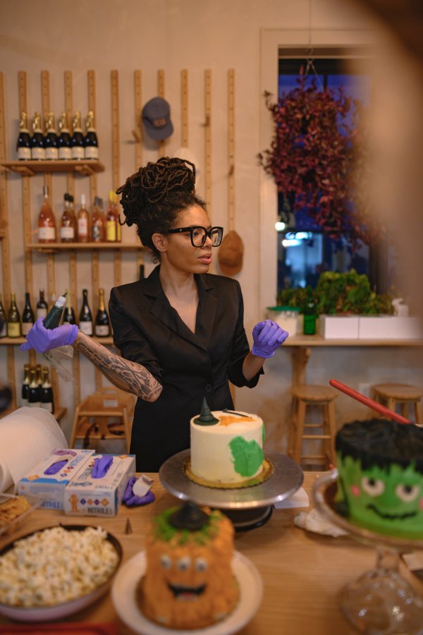 A woman with dreadlocks, glasses, and tattoos wearing purple gloves, decorating a cake in a cozy kitchen with wine bottles and herb plants, preparing for a celebration.