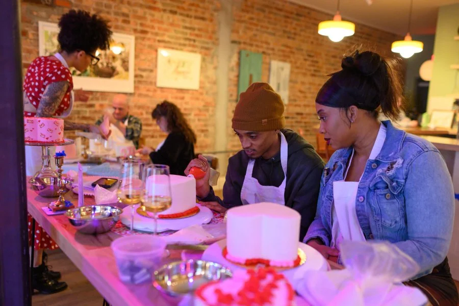 People decorating cakes at a cooking class or workshop in a cozy, brick-walled kitchen or studio.