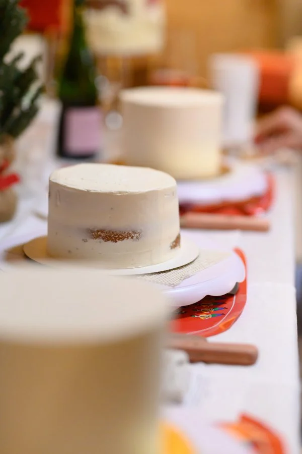 Two white frosted birthday cakes on a table, with one cake in focus in the foreground and the other in the background, surrounded by festive holiday decorations.