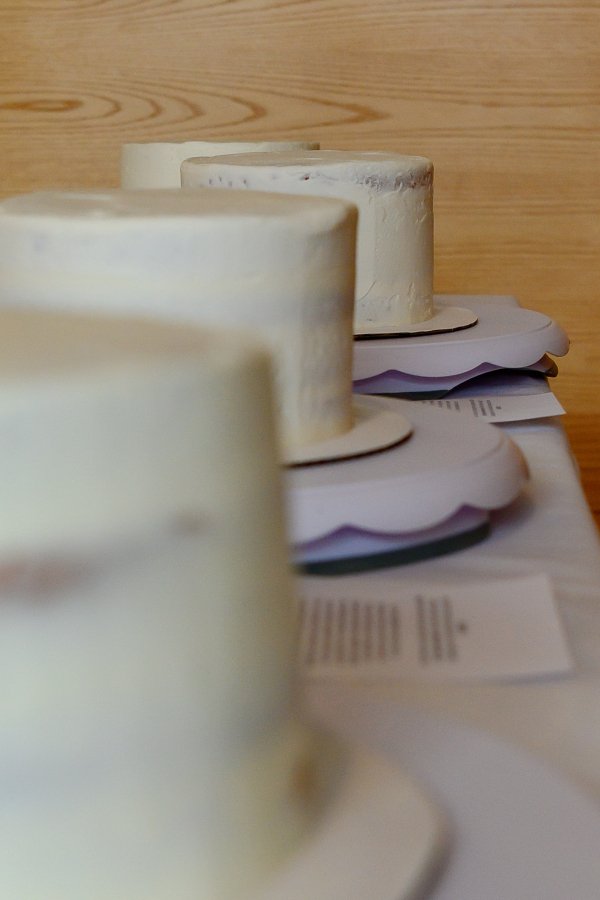 Multiple white frosted cakes on wooden tables, with sheets of paper and other items underneath, in a row.