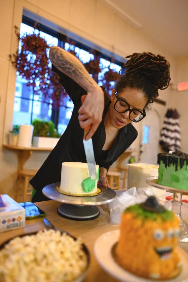 A woman with glasses and dreadlocks decorates a cake with green frosting in a cozy kitchen. There are other decorated cakes and food items on the table, with autumn leaves visible outside the window.