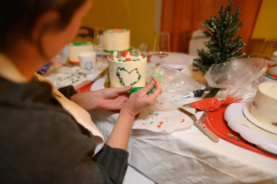 A person decorating a Christmas cake with green icing, surrounded by other cakes, a small Christmas tree, and decorating tools on a table.