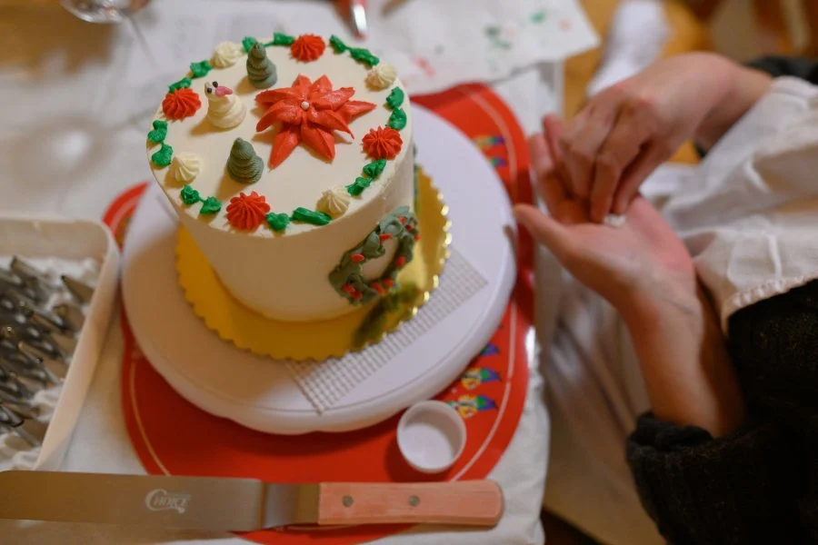 A Christmas-themed cake decorated with poinsettias, Christmas trees, and other holiday motifs on top. The cake is on a white stand, and a person is holding hands with another person nearby.