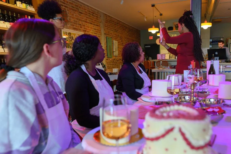 Group of women in a restaurant, one woman standing and taking a photo with a phone while others sit at a table with wedding cakes and glasses of wine or champagne.