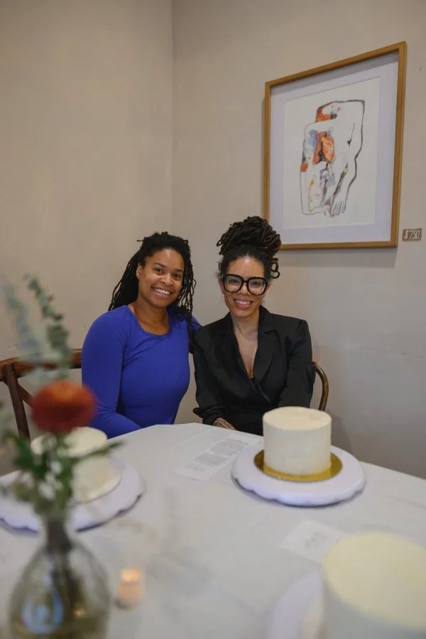 Two women sitting at a table with a white cake in front of them, smiling at the camera. One woman is wearing a blue top and the other is wearing a black blouse and glasses. A framed abstract artwork hangs on the wall behind them.