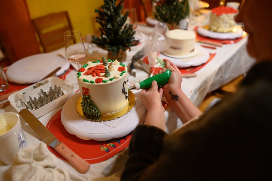 Person decorating a holiday cake with a piping bag at a Christmas dinner table with small Christmas trees and other desserts.